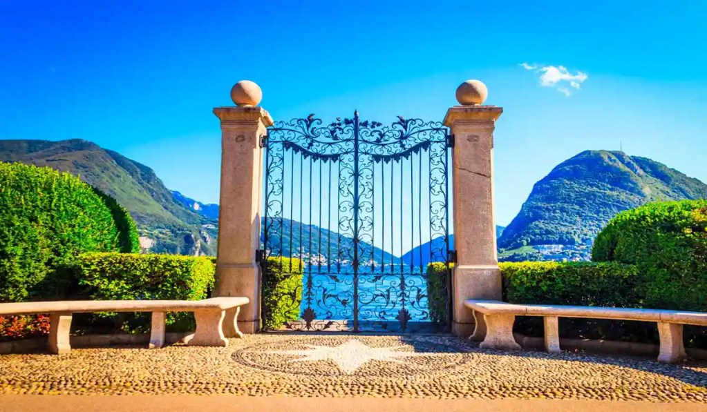 An elegant wrought-iron gate in Parco Ciani, Lugano, Switzerland, framed by stone pillars and lush greenery, opens to reveal a stunning view of Lake Lugano and the surrounding mountains. Two stone benches sit on either side of the gate, offering a peaceful spot to enjoy the picturesque scenery under a clear blue sky.