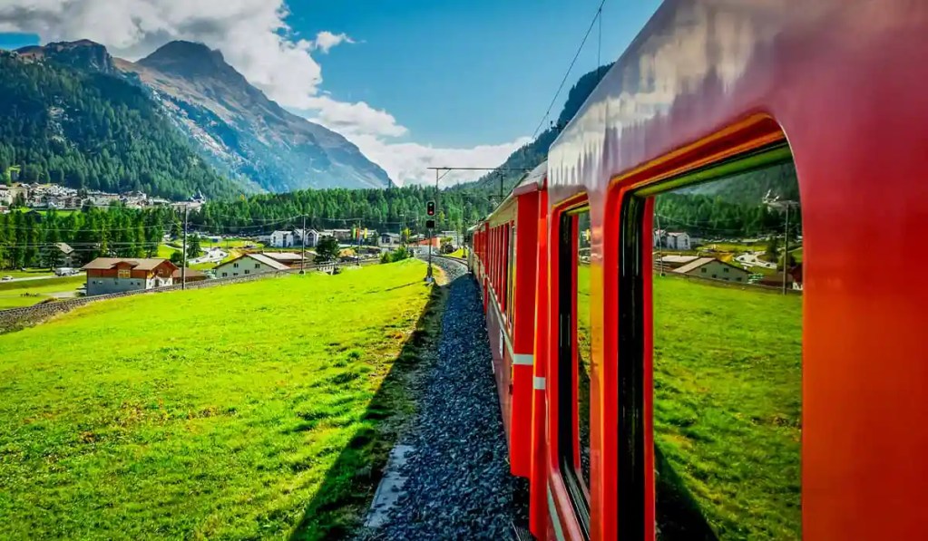 A picturesque journey on a red train traveling through the Swiss countryside, showcasing lush green fields, quaint houses, and forested hills. Majestic mountains tower in the background under a partly cloudy sky, capturing the scenic beauty of Switzerland along the route from Lugano to Zurich.