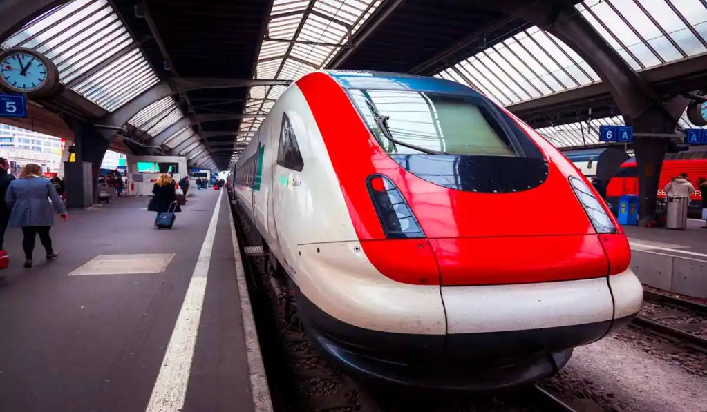 A sleek, modern Swiss train parked at a station platform under a glass-roofed structure, with passengers walking alongside and boarding. The train's red and white design contrasts with the station's industrial architecture, creating a dynamic and bustling travel scene.