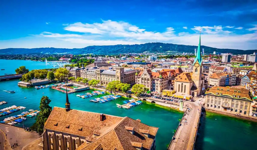 This vibrant image captures the picturesque cityscape of Zurich, Switzerland, with a focus on the Fraumรผnster church and its distinctive green spire. The scene includes the turquoise waters of the Limmat River, numerous boats docked along the shore, and the backdrop of the lush, rolling hills.