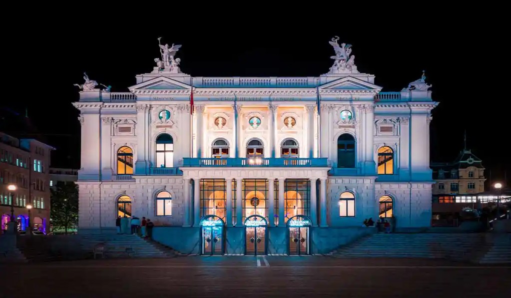 This image showcases the Zurich Opera House illuminated beautifully at night. The grand neoclassical facade is adorned with statues and columns, with the building's lights casting a warm glow, highlighting its architectural elegance.