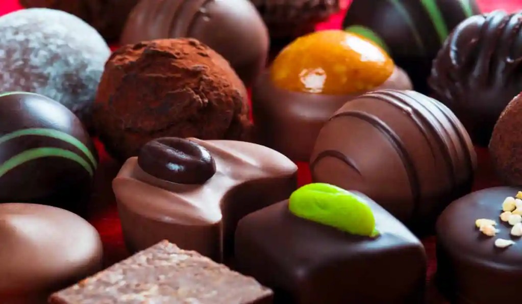 This close-up image features an assortment of Swiss chocolates, showcasing various truffles and pralines with different textures and toppings. The chocolates are decorated with ingredients like cocoa powder, nuts, coffee beans, and vibrant green accents, highlighting their rich and indulgent appearance.