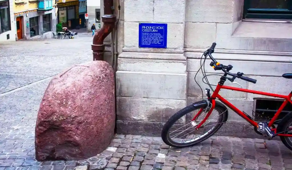 This image features the Findling vom Geissturm, a historical stone landmark in Zurich, Switzerland. A red bicycle is parked next to the stone, which is situated against a building with a blue informational plaque. The cobblestone street and surrounding buildings create a quaint, European cityscape atmosphere.