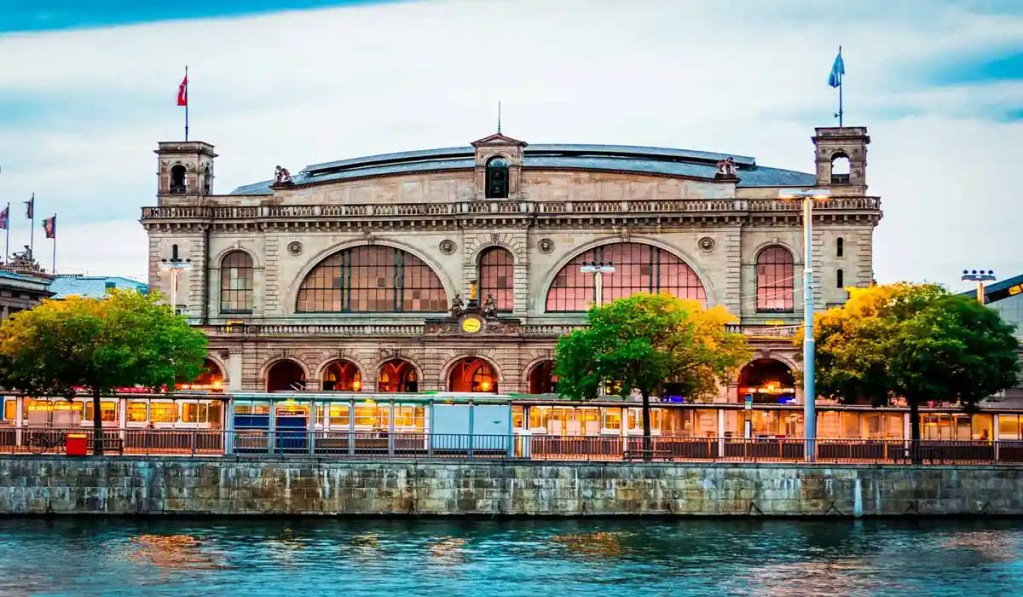 A view of the grand Zurich Hauptbahnhof (main train station) in Zurich, Switzerland, showcasing its impressive facade with large arched windows and classical architectural details. The station is situated along a river, with trees and a tram adding to the bustling urban atmosphere under a slightly overcast sky.