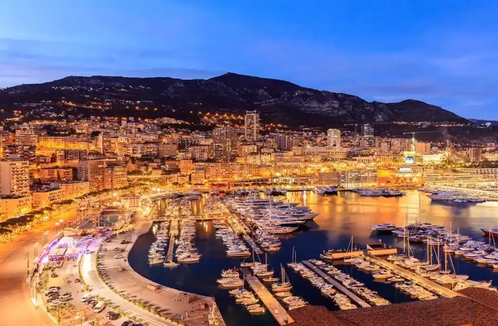 A stunning nighttime view of Monaco’s coastline, showcasing a marina filled with luxury yachts. The city is illuminated with warm lights, casting a golden glow over the buildings and reflecting on the water. In the background, rolling hills and mountains rise up, dotted with lights from the surrounding residences. The contrast between the vibrant cityscape and the calm harbor creates an inviting and luxurious atmosphere.