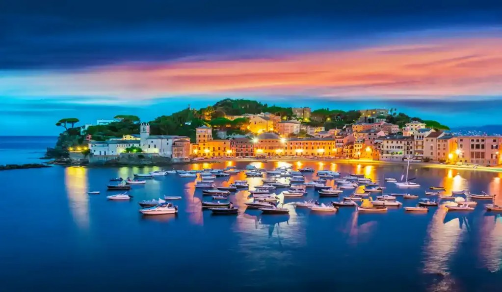 This image captures the enchanting Bay of Silence (Baia del Silenzio) in Sestri Levante, Italy, during twilight. The bay is illuminated by warm lights from the surrounding colorful buildings, while numerous small boats float peacefully on the calm water, reflecting the vibrant hues of the sunset and the deep blue sky.