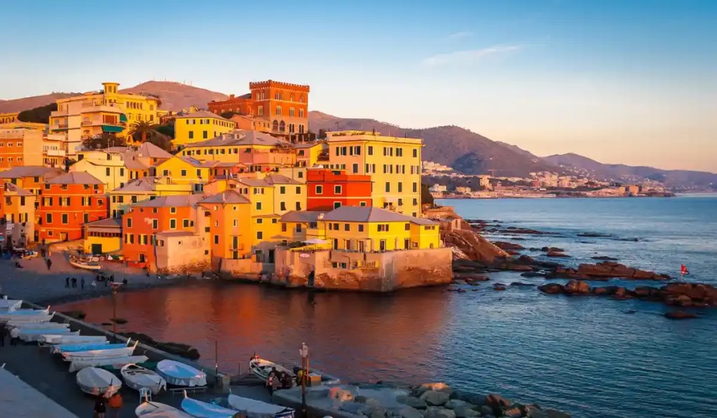 This image shows the picturesque coastal village of Boccadasse in Genoa, Italy, during golden hour. Vibrant pastel-colored buildings line the rocky shore, reflecting on the calm waters, with boats resting on the promenade and distant hills completing the serene scene.