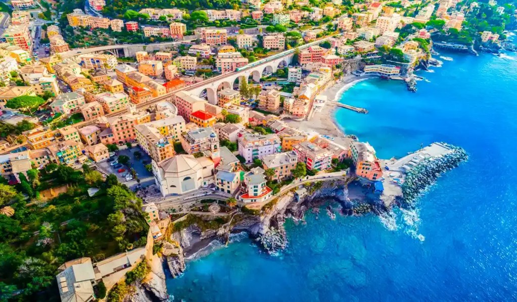 This aerial image captures a vibrant coastal town near Genoa, Italy, showcasing its colorful buildings, curved bay, and turquoise waters lapping on the shore of Spiaggia Libera Sotto La Chiesa. The town is framed by lush greenery and features a prominent viaduct running through the landscape, connecting its charming neighborhoods.