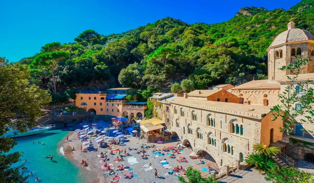 This image captures the secluded beach and historic Abbey of San Fruttuoso, nestled along the Ligurian coastline in Italy. Surrounded by lush green hills, the abbey's stone architecture overlooks a small, pebbly beach filled with colorful umbrellas and sunbathers, while turquoise waters lap the shore.