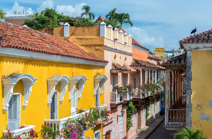 Charming colonial street scene with brightly painted yellow and pastel buildings, white-trimmed balconies overflowing with colorful flowers, red clay tile roofs, and palm trees peeking over the rooftops under a clear blue sky.