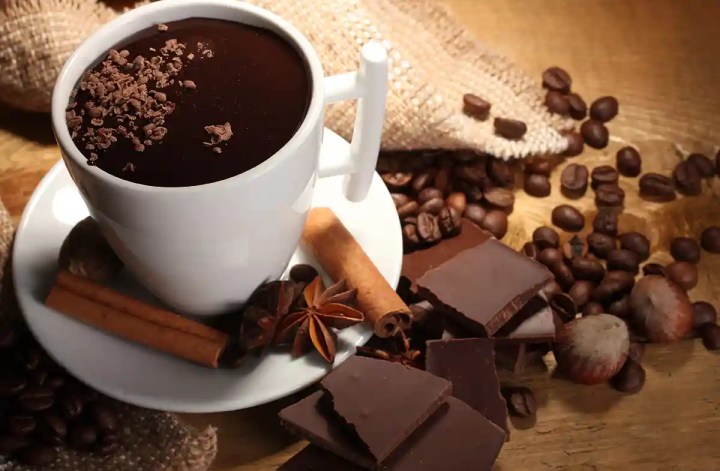 A white mug filled with rich hot coffee, surrounded by cinnamon sticks, star anise, dark chocolate chunks, coffee beans, and a burlap sack on a wooden surface.
