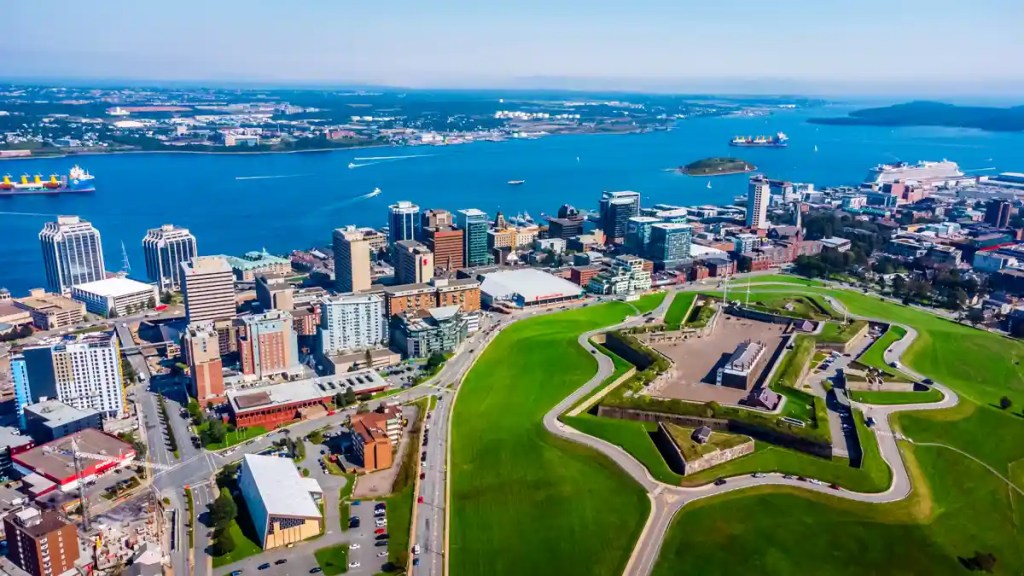 Aerial view of downtown Halifax, Nova Scotia, highlighting the star-shaped Halifax Citadel National Historic Site surrounded by green lawns, with the harbor and waterfront skyline in the background, and ships visible in the blue waters of the harbor.